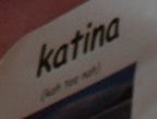 Close-up of a sign with the word "katina" in bold black text. The background is blurred with soft, muted colors.