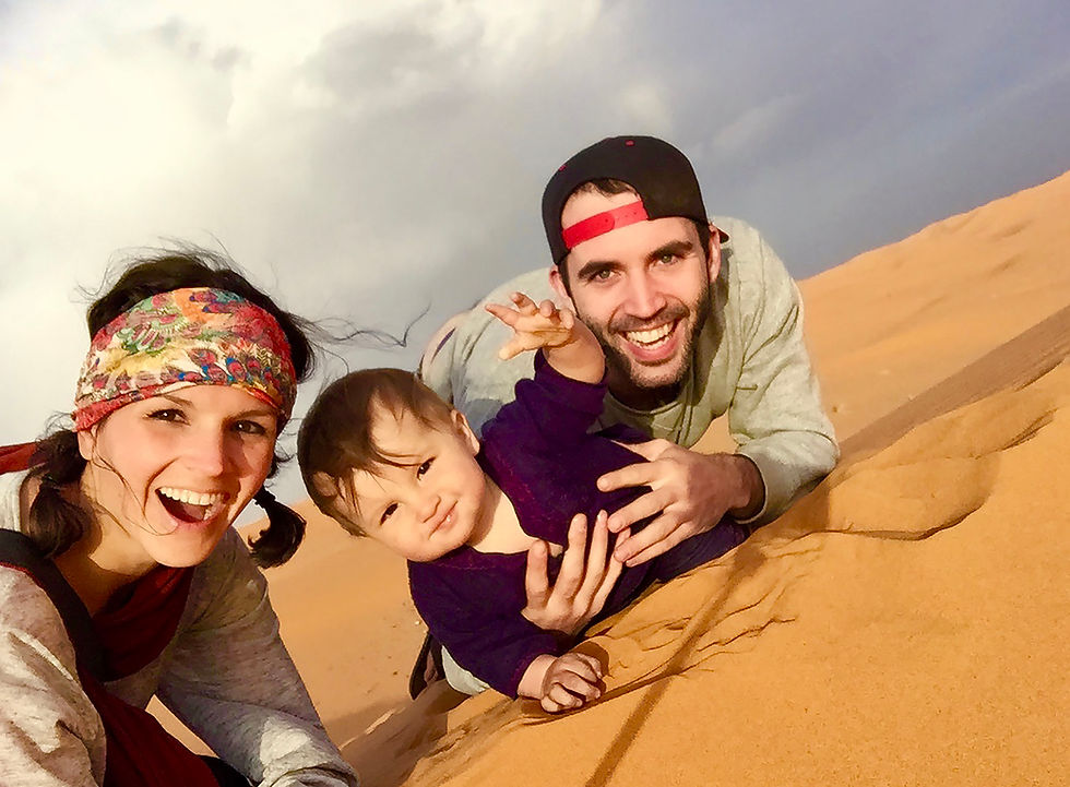Smiling couple and baby lying on sandy desert. Woman wears a colorful headband, baby in purple, man in a cap. Cloudy sky backdrop.