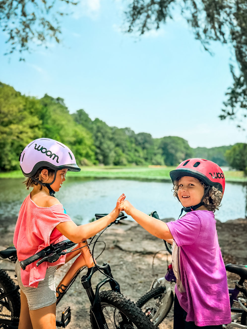 Two kids in pink Woom helmets high-five by a lake, smiling. They're wearing bright shirts and standing with bicycles on a sunny day.