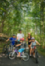 Family of five biking on a forest trail, smiling under leafy trees. Bright helmets and casual attire enhance the joyful outdoor vibe.