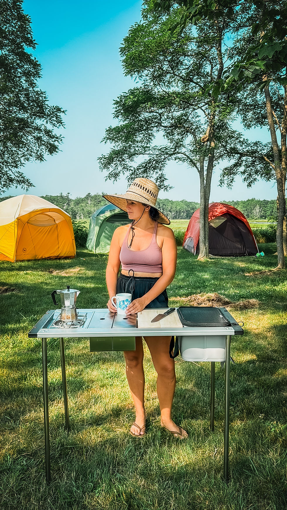 Woman in a hat holding a mug at a campsite table with a coffee maker. Tents and trees in the background, bright and sunny setting.