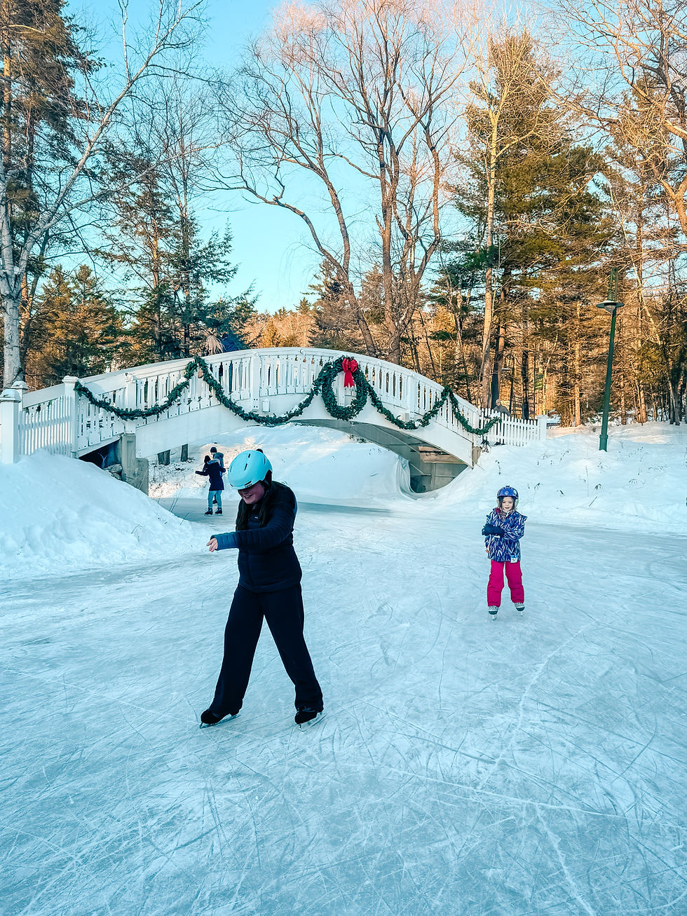 Two people ice skating on a snowy rink in North Conway NH. A festive bridge with garlands is in the background. Bright winter clothing, clear blue sky.