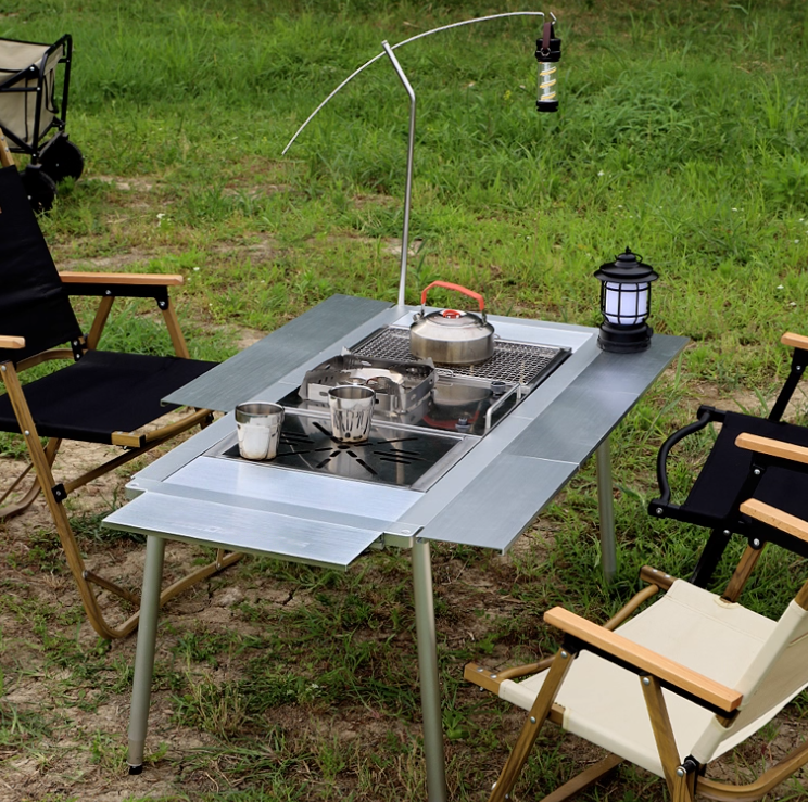 Thumbnail: キャンプテーブルと調理器具。金属テーブルに料理、ランタン、キャンプ椅子、背景は緑地。