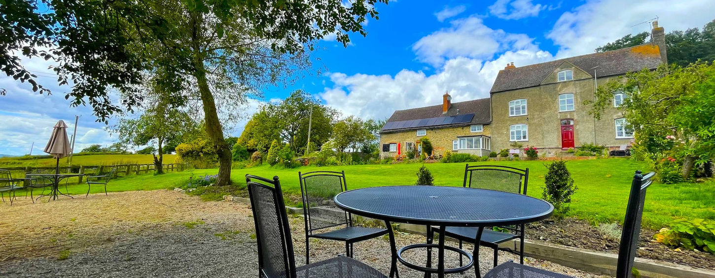 Gravel seating area with table and chairs at The Wellspring Ledbury, overlooking lawn, trees, and main house and cottage in the background