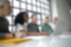 A focused group of professionals sitting at a white table, collaborating and looking intently at a laptop screen in a bright, industrial-style room with large windows.