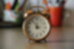 Close-up of a vintage-style alarm clock showing the time as 6:00, placed on a wooden surface with blurred background items.
