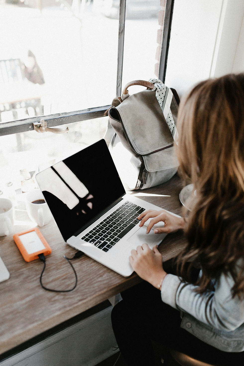 A woman sits at a wooden desk by a window, working on an open laptop. A gray backpack is resting on the chair, and a bright orange external hard drive is connected to the laptop. The workspace looks cozy with a cup of coffee nearby.