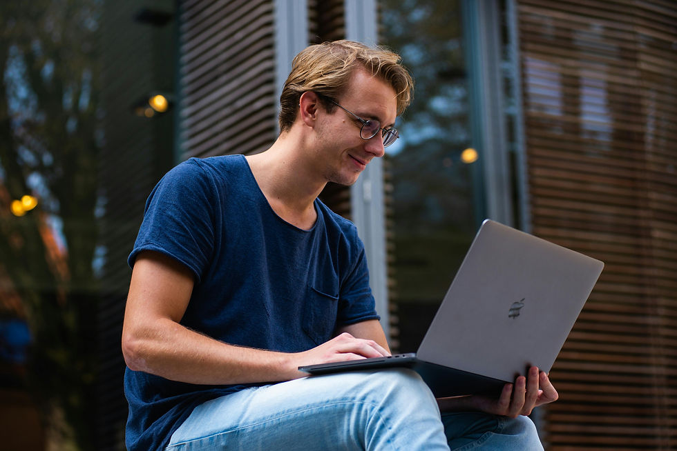 A smiling man sitting outdoors while working on a laptop. He appears to be relaxed and concentrated, possibly working remotely.