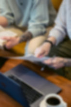 Two people reviewing printed documents while working on a laptop, with a cup of coffee nearby, suggesting a focused study or work session.