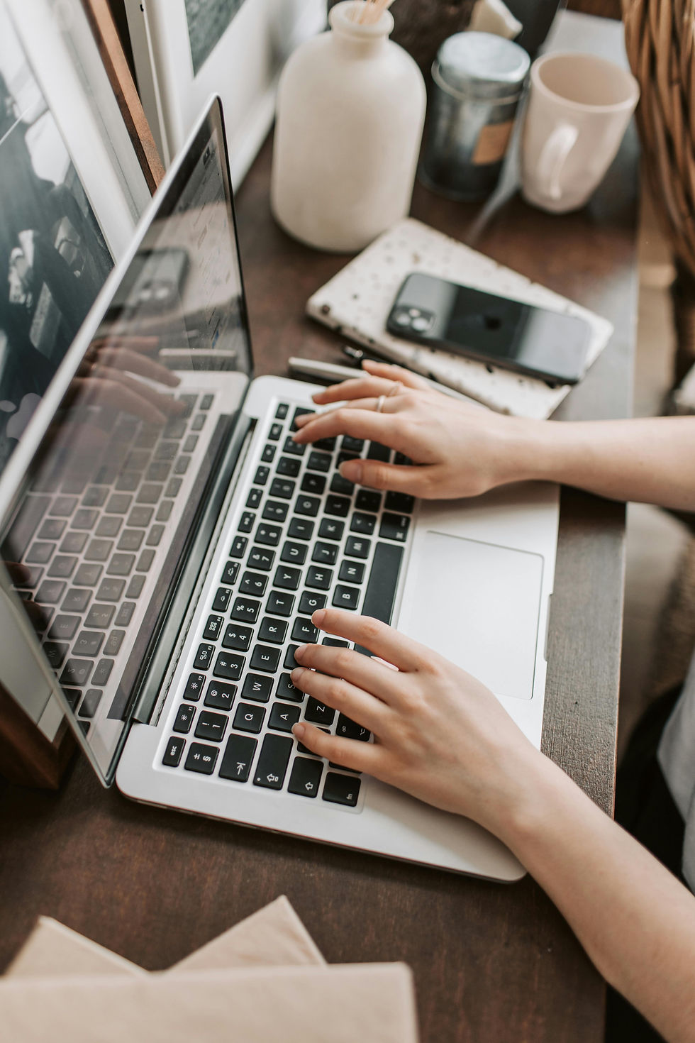Close-up of a person's hands typing on a laptop keyboard, with a smartphone and a notebook beside them.