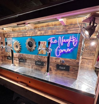 The side of a beer tap truck shows four silver bar taps, 3 small vintage gold mirrors hand decoravtove as the backdrop, along with a pink neon sign reading 'the naughty corner' and some vintage alcohol tin signs