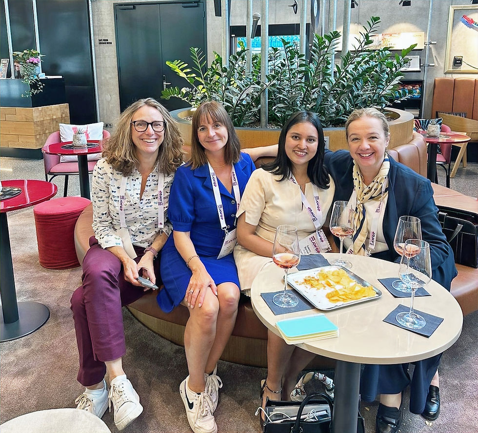 Four women smiling, sit on a couch with drinks and snacks on a table. They're in a modern lounge with plants, wearing conference lanyards.