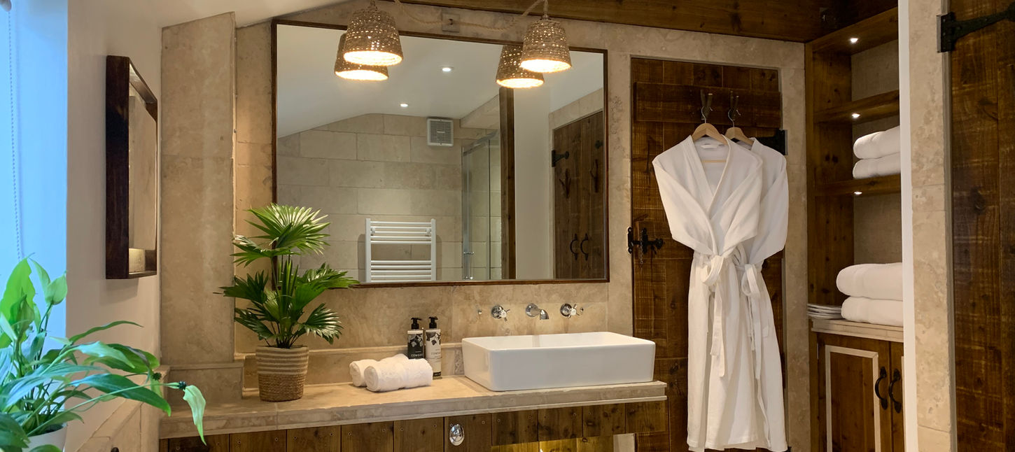 Warm, spa-like bathroom with stone walls, wooden accents, a white robe, plants, and neatly folded towels at Dovecote Cotswold Cottages