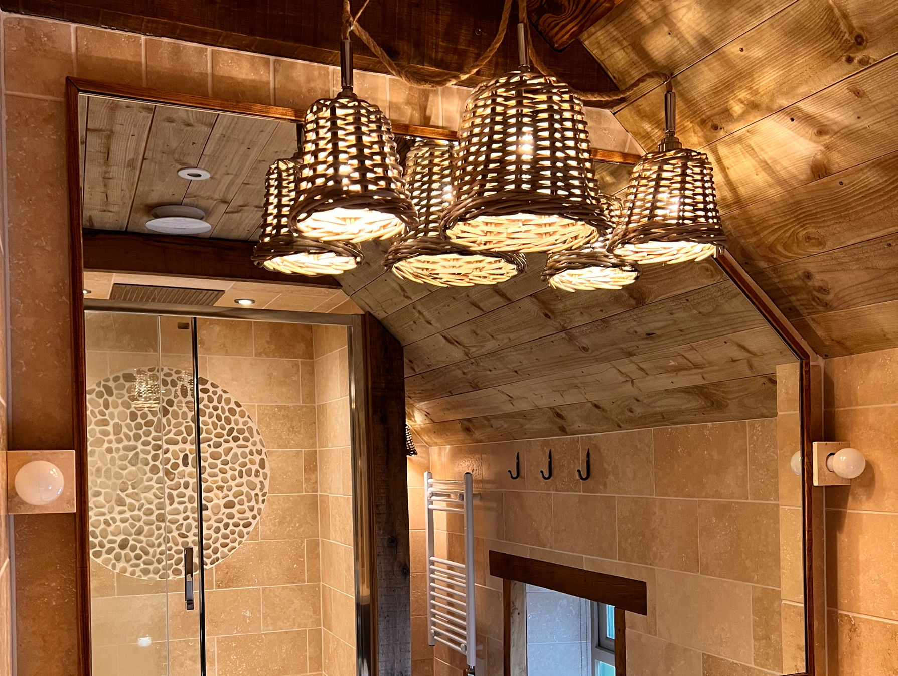 High-spec bathroom in a Cotswold holiday rental showing rustic wooden ceiling beams, basket-weave light fixtures, and travertine wall tiles.
