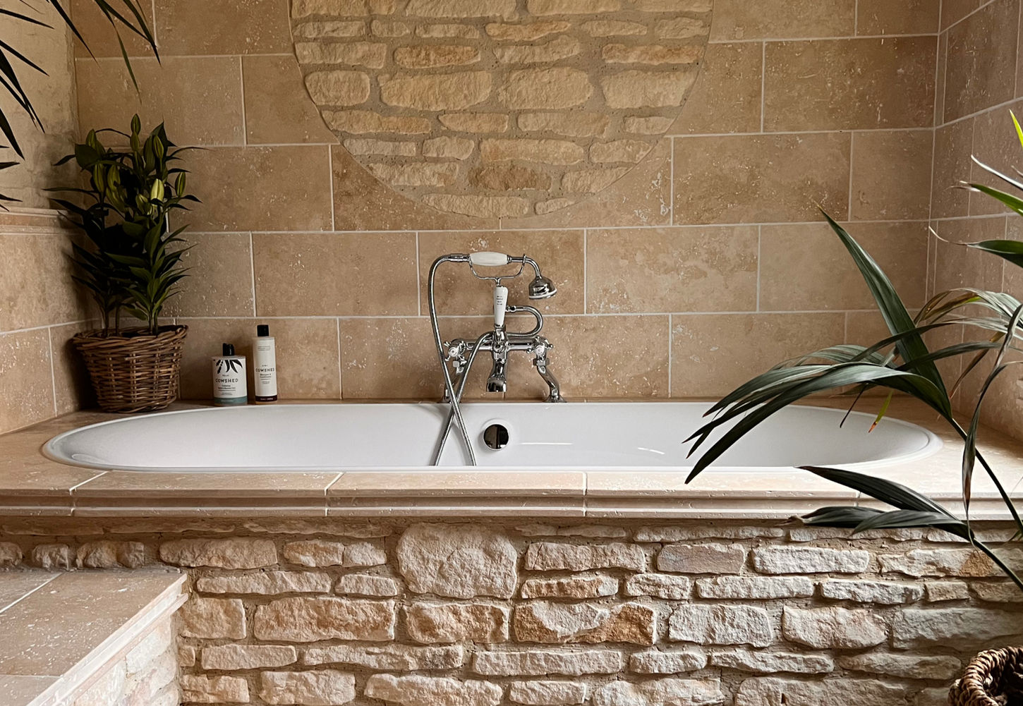 Stone-lined bathtub with traditional chrome tap, soft lighting and surrounding plants at Dovecote Cotswold Cottages. Cowshed bath and shower gel, Cowshed shampoo and Cowshed hand wash.