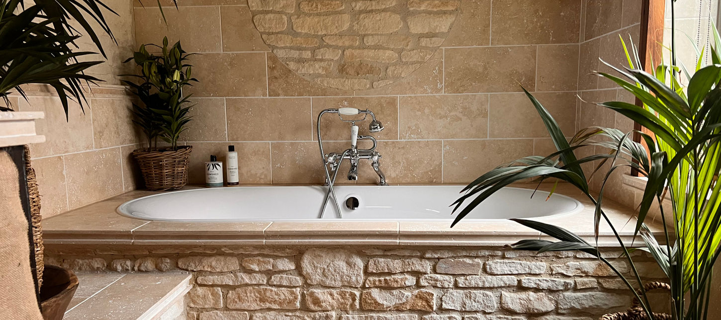 Stone-lined bathtub with traditional chrome tap, soft lighting and surrounding plants at Dovecote Cotswold Cottages. Cowshed bath and shower gel, Cowshed shampoo  and Cowshed hand wash.
