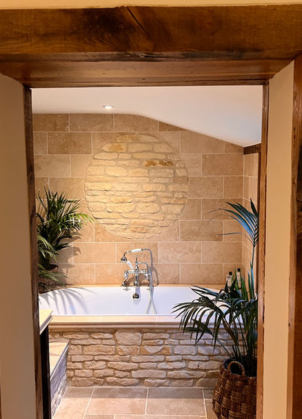 A gorgeous and luxurious bathroom with palms surrounding it. The large bath is surrounded by a hand-crafted wall made of Cotswold stone and the walls of the bathroom are also made of stone. This makes the room feel relaxing, a perfect place to unwind.