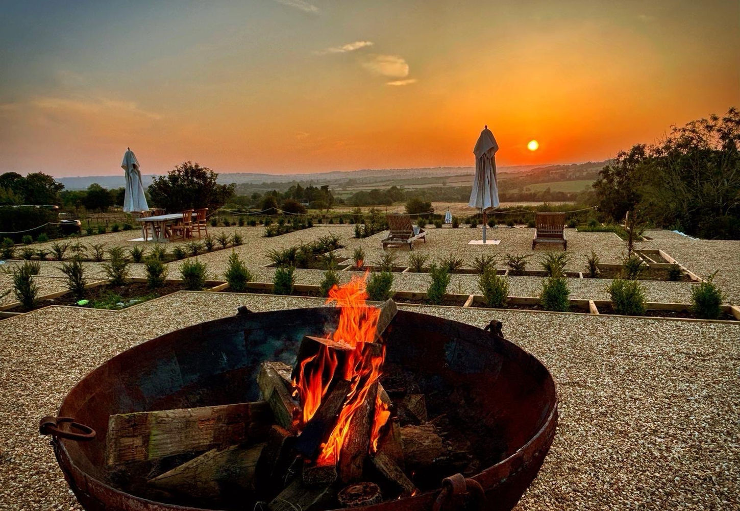 Sunset over the Cotswold countryside with a fire pit, outdoor seating, and landscaped gardens at Dovecote Cotswold Cottages.Valley. A fire-pit and parasols in view.