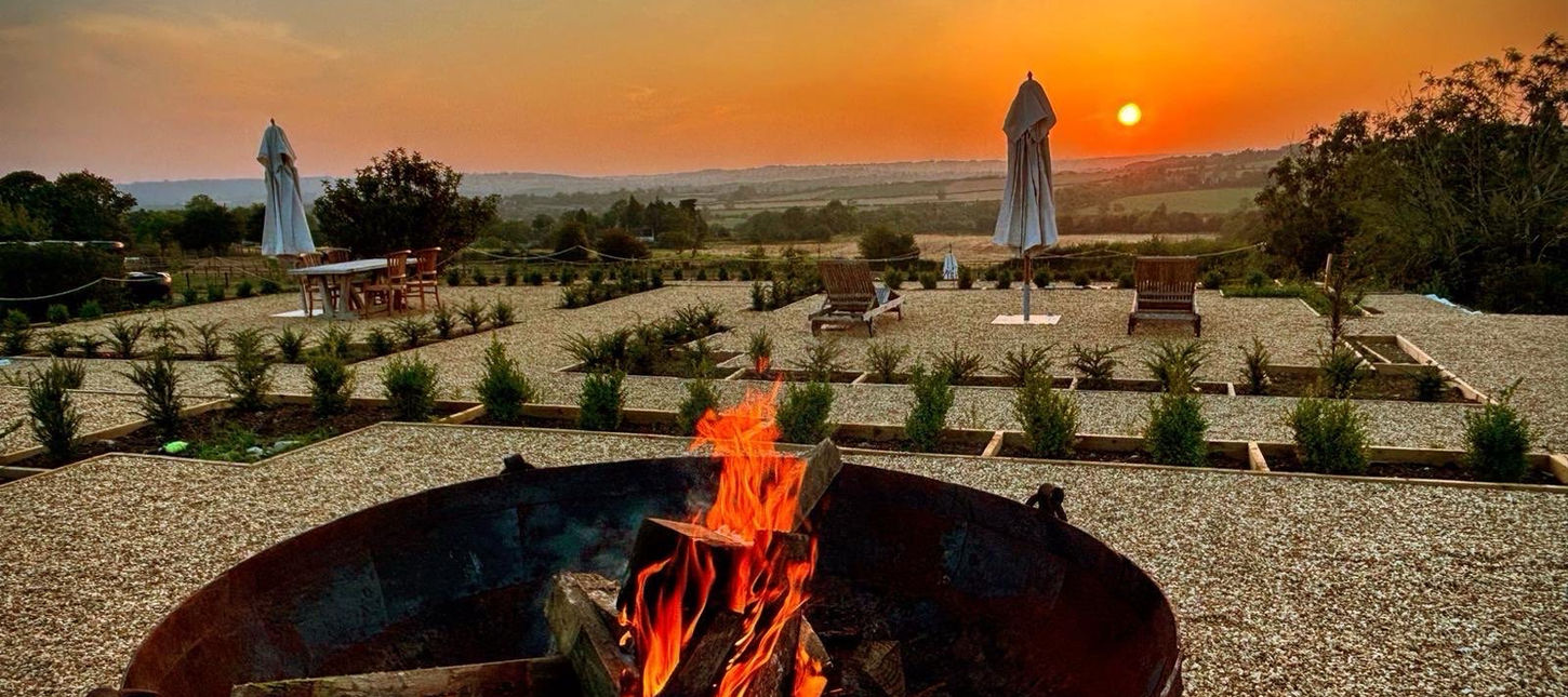 Sunset over the Cotswold countryside with a fire pit, outdoor seating, and landscaped gardens at Dovecote Cotswold Cottages.Valley. A fire-pit and parasols in view.