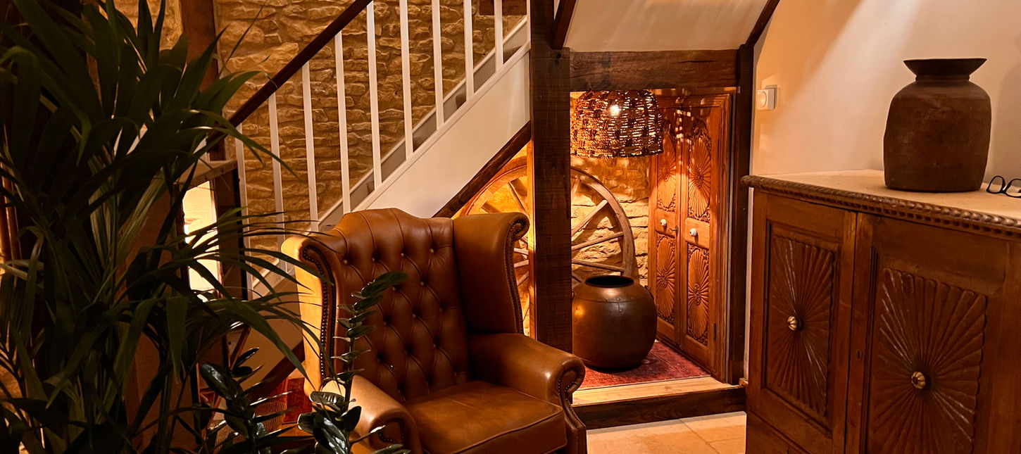 Warmly lit interior with leather armchair, carved wooden furniture, stone walls and rustic decorative elements at Dovecote Cotswold Cottages