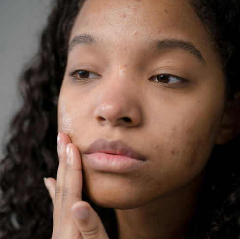 A brown skinned woman with dark hair spreads skin care product on her cheek