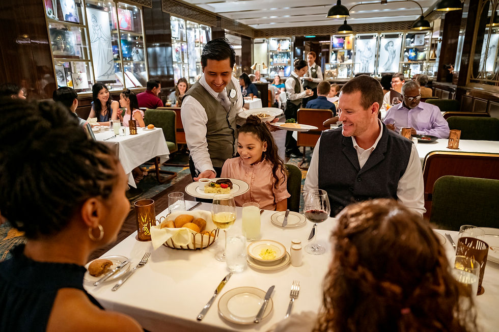 Server presents a dish to a smiling child at a dining table. Others look on in a warmly lit restaurant. Art adorns the walls.