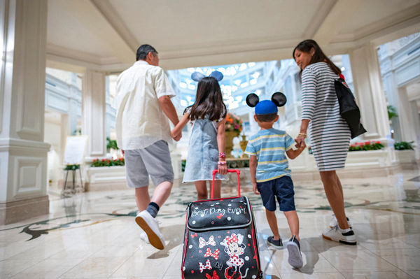 Family of four in a hotel lobby, children wearing Mickey hats. A bag reads "Best Day Ever!" Mood is joyful with muted colors.