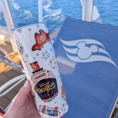 Hand holding a white tumbler with Disney Destiny text and ship graphic, against a blue ocean backdrop with a lounge chair featuring a Mickey logo.