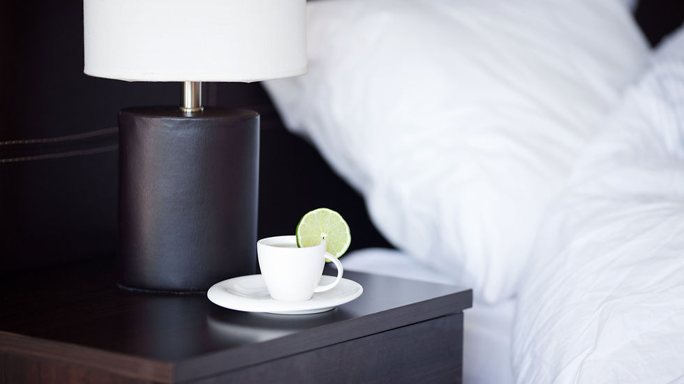 White cup with a lime slice on a dark bedside table next to a lamp and fluffy white pillows. Peaceful and minimalist setting.