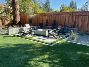 Backyard patio with synthetic turf and modern concrete fire pit surrounded by Adirondack chairs.