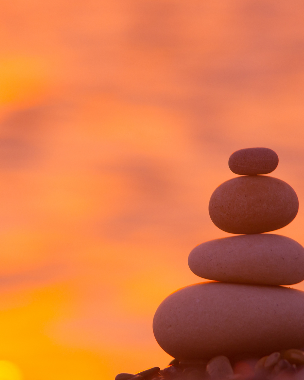 Four smooth rocks stacked on top of each other, each smaller than the one under it, with a calming sunset backdrop, representing how busy women can finish the year in peace with a year-end reflection.