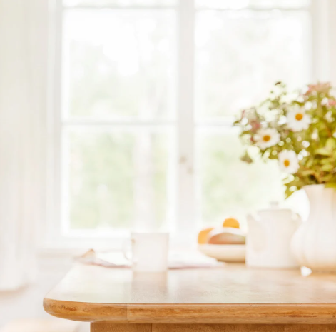 A partial photo of a dining table, clear of any clutter, with only a tea pot, a mug, a flower vase, and a book. The sun is shining through the window setting a clam scene, representing the mental clarity that can be achieved with a simple home organization routine.