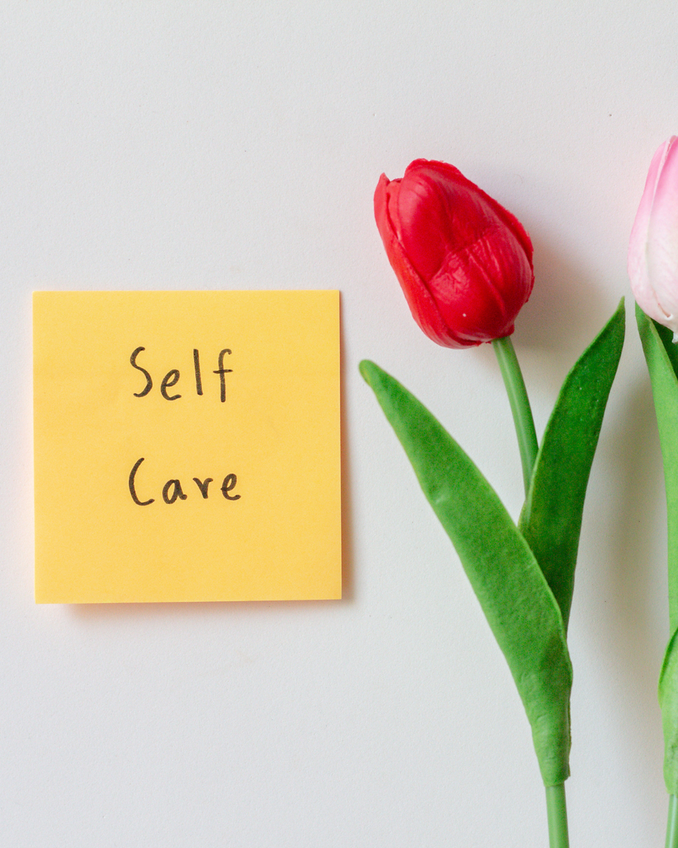 A tulip in front of a blank wall with a sticky note reading "Self Care," representing the importance of daily resets as a form of stress relief for busy moms.