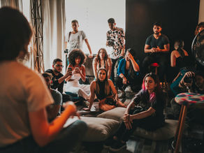 A group of people sit attentively on cushions indoors, some taking photos. The room is dimly lit, with a casual and engaged atmosphere.