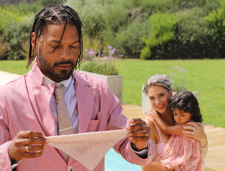 A man in a pink suit holding a cloth by a poolside, with a woman in a glittery pink dress carrying a child in matching attire
