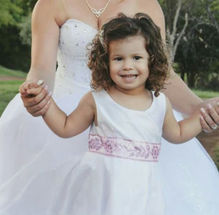 Close-up of a little girl wearing a white sleeveless dress that has a pink waist detail. The bride is holding her hands.