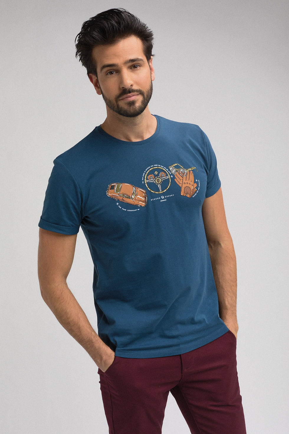 Photo couleur de mode masuline. Un homme porte un tshirt de la marque "Benson & Cherry" de couleur bleu avec une sérigraphie représentant une voiture, un volant, une paire de gants ainsi qu'une paire de lunette. L'homme sur la photo est de face et a un regard franc et souriant. La photo est cadrée en format portrait.