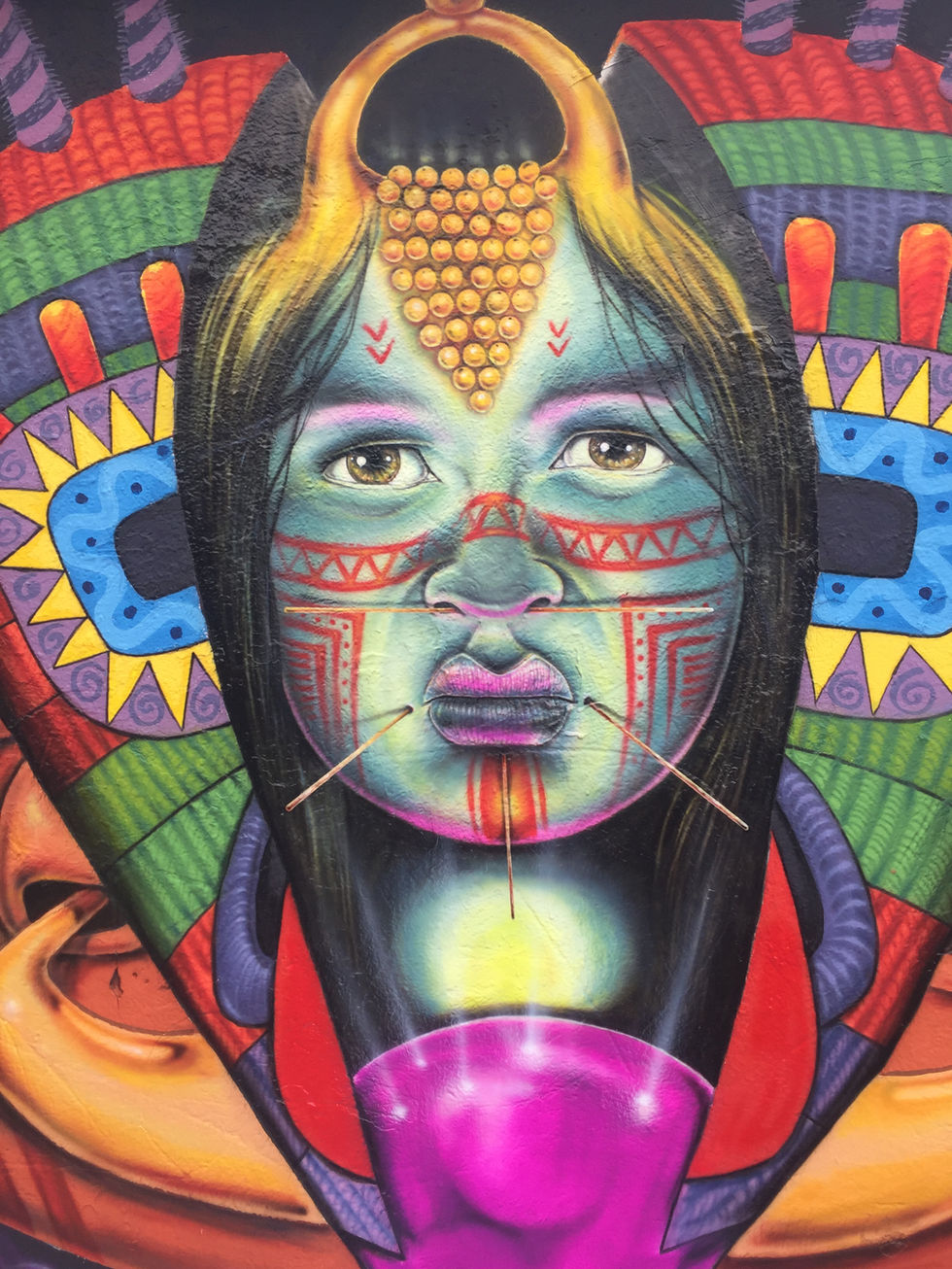 Primer plano de detalle de la sección central de un mural de arte público. Muestra el rostro estilizado de una mujer indígena en tonos turquesas, con pintura facial tradicional en rojo y negro, palillos ornamentales de madera atravesando el labio y la nariz, y un tocado dorado sacerdotal sobre la frente.