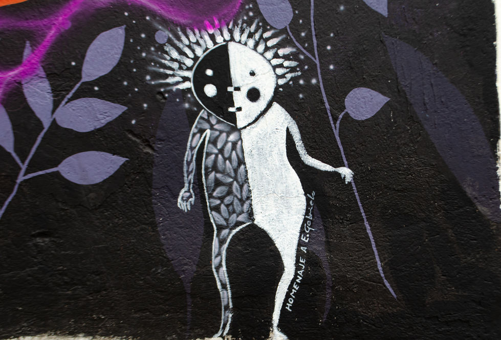 Fotografía de detalle en la base inferior del mural Arqueología del Ser (2026). Muestra una esfera violeta central flanqueada por pequeñas figuras lineales y etéreas de color blanco y negro en actitud ritual. Este segmento constituye un homenaje plástica y una cita visual conmemorativa a la fallecida muralista urbana Gabriela Ayala (1973-2023), desarrollado mediante una hibridación técnica asistida por Inteligencia Artificial y ejecutada en aerografía sobre el muro institucional.