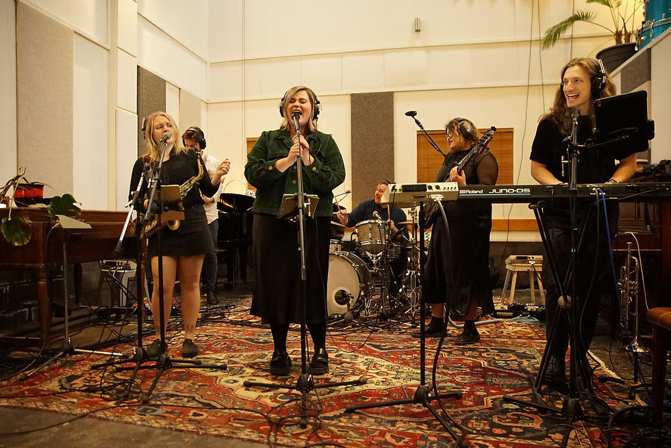 Band performing in a studio; singers and musicians with instruments on a vibrant rug. Headphones worn, lively and engaging atmosphere.