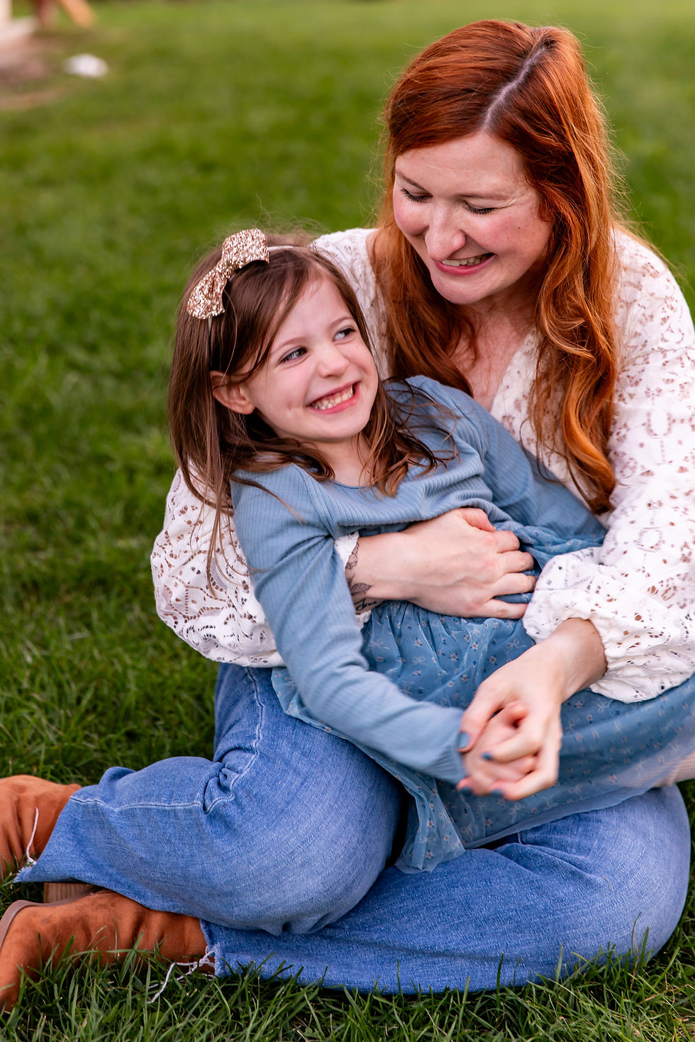Woman with red hair hugs smiling girl in blue dress on grass. Both appear happy. Woman wears lace top, girl has sparkly headband.
