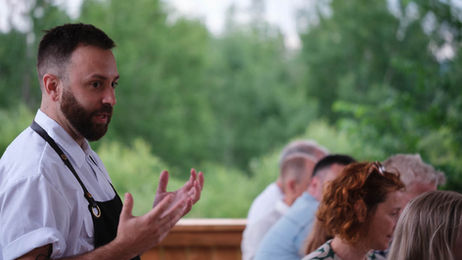 Chef montréalais en tablier noir s’adressant aux convives lors d’un souper champêtre aux Cocagnes à Frelighsburg, avec un fond de verdure estivale et une ambiance conviviale