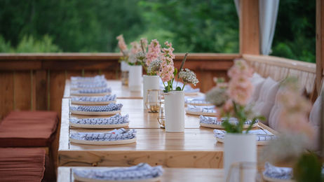 Table en bois dressée sous une pergola aux Cocagnes, avec vaisselle soignée, serviettes à motifs, fleurs fraîches en centre de table et bougies, prête à accueillir les convives pour un souper champêtre à Frelighsburg.