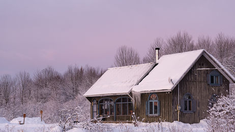 Maison en bois recouverte de neige au site agroécologique Les Cocagnes à Frelighsburg, entourée d’un paysage hivernal sous un ciel rosé en fin de journée."