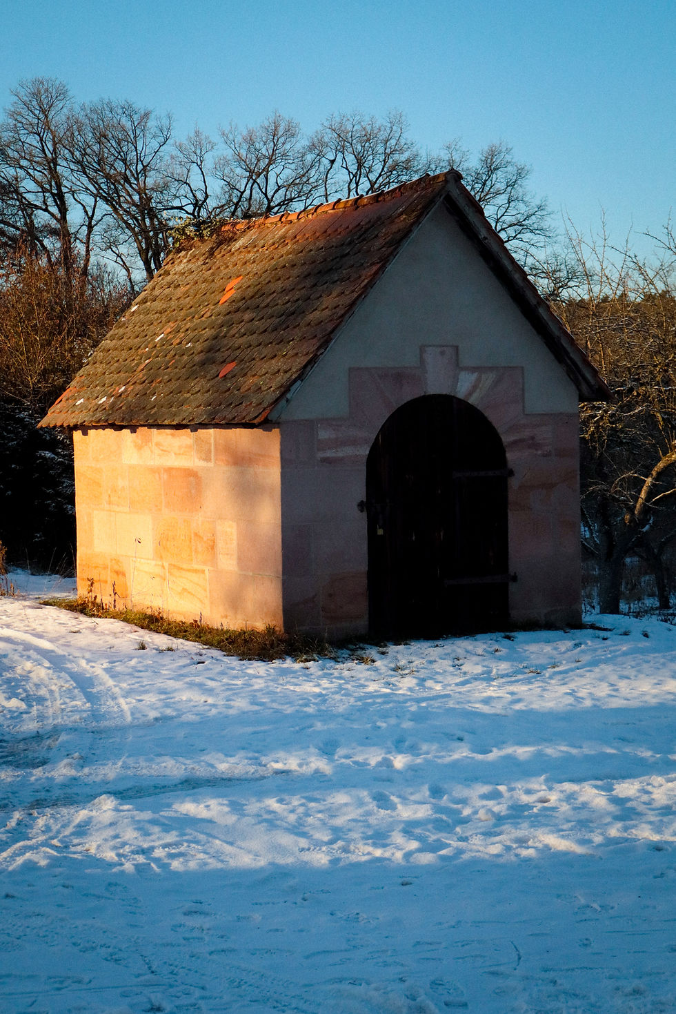 Small stone shed with a tiled roof in a snowy landscape. Bare trees and clear blue sky in the background. In front of is there's a snow on the ground, shadows apearing blue.