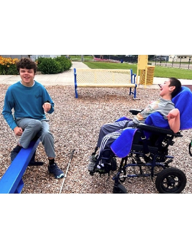 A young man in a wheelchair laughs while sitting beside another young man on a teeter-totter, both smiling