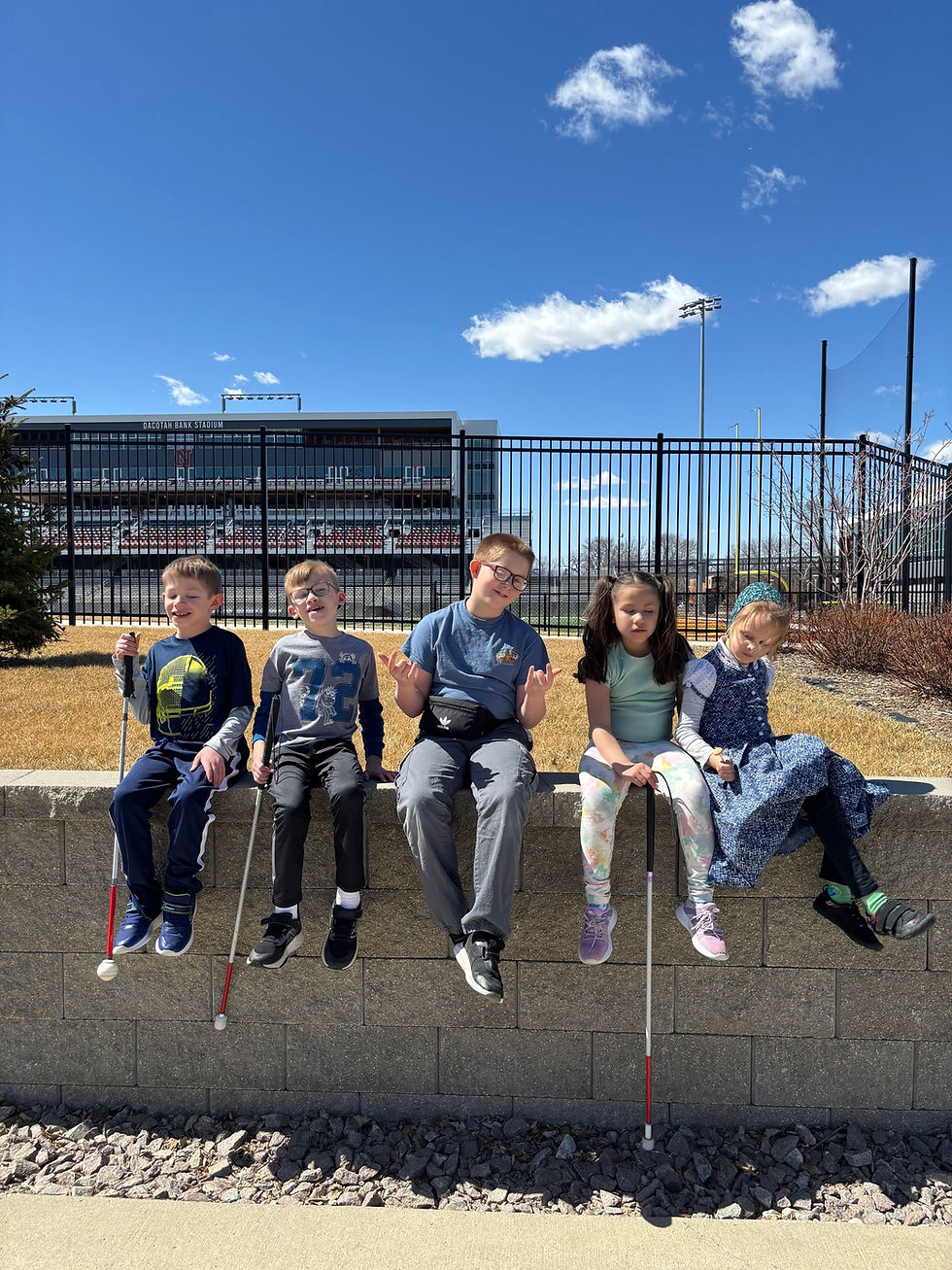 A group of five young students sitting in a line on top of a brick wall in front of Dacotah Bank Stadium on the campus of Northern State University