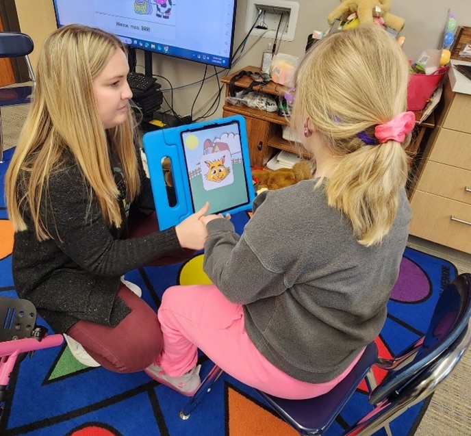 A woman kneels in front of a young girl, holding an iPad as they interact_