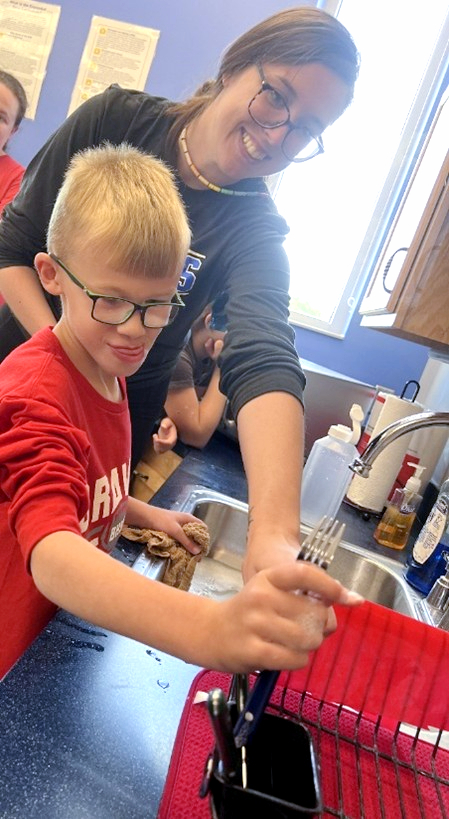 A young boy stands at a sink filled with soapy water, placing a fork into a dish rack_