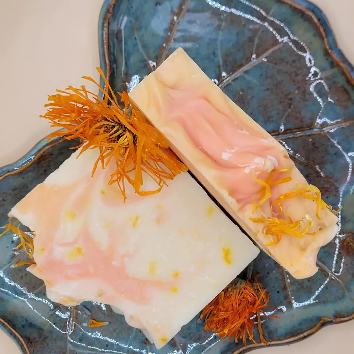 dwie kostki mydła nagietkowego w otoczeniu suszonych kwiatów na ozdobnym ceramicznym talerzyku